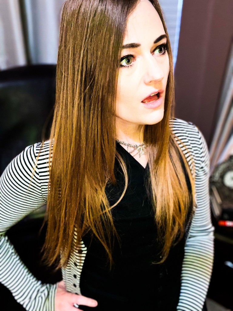 A person with long, straight hair, wearing a striped cardigan, looking surprised or shocked while standing indoors.