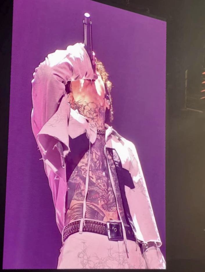 Close-up of a male singer performing on stage, holding a microphone, with visible tattoos and a dynamic pose against a purple background.