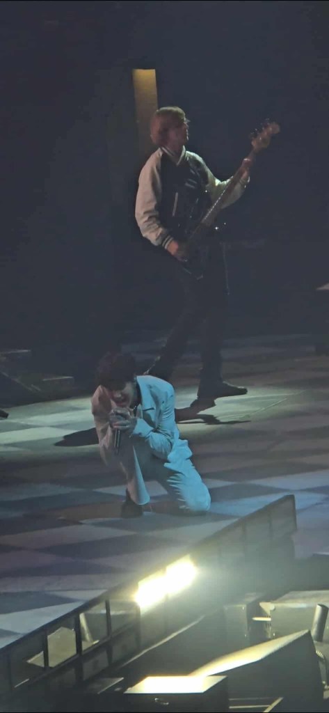 A live concert image featuring a vocalist kneeling on stage, passionately singing into a microphone while a guitarist stands in the background, illuminated by stage lights.