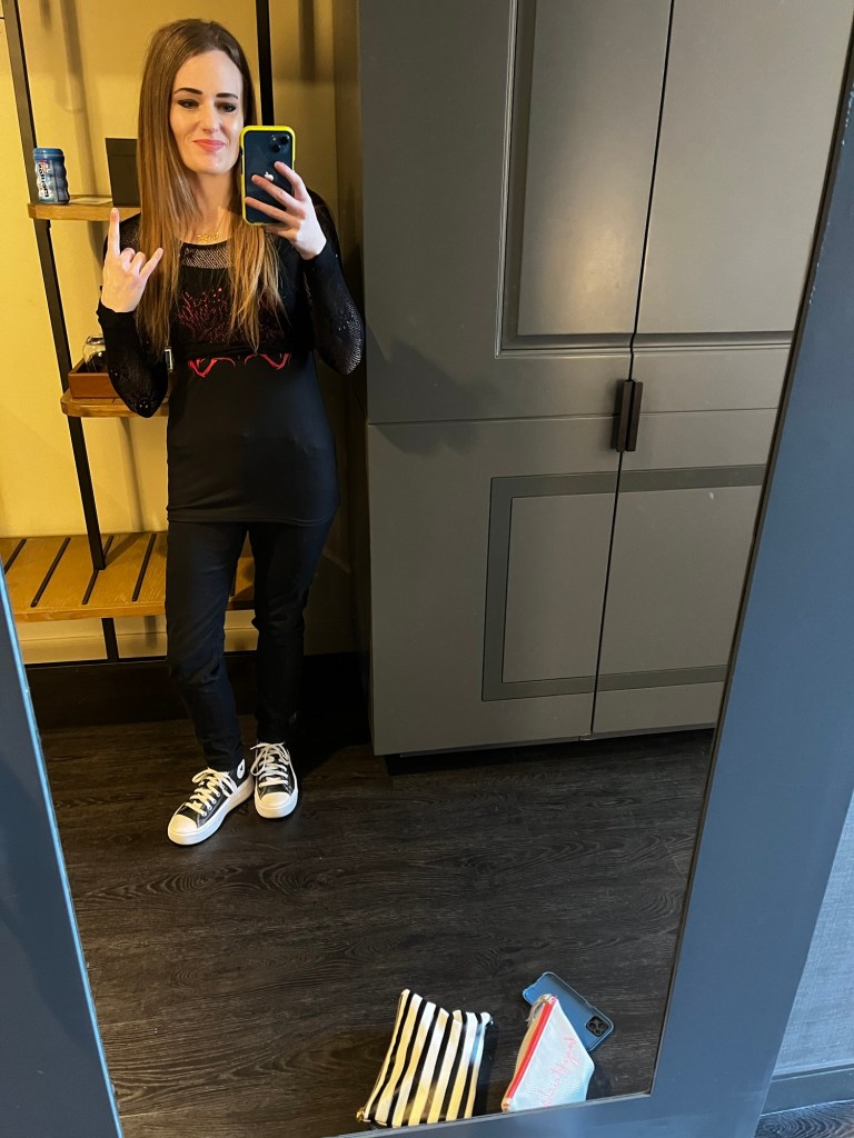 A person taking a selfie in a mirror, wearing a black outfit with rock-themed graphics, showing a hand sign, and standing in a stylish room with wooden shelves.