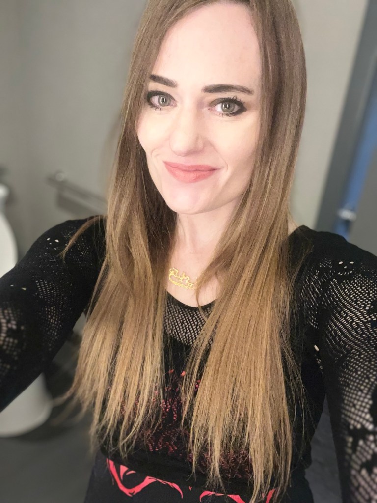A smiling individual with long brown hair is posing for a selfie, wearing a black mesh top and a necklace, with a subtle background that includes a bathroom setting.