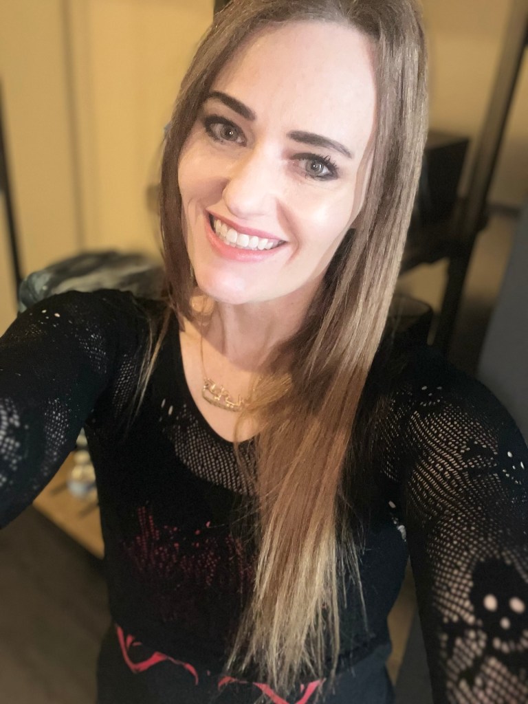 A smiling woman with long hair wearing a black top with a lace pattern, holding the camera for a selfie.