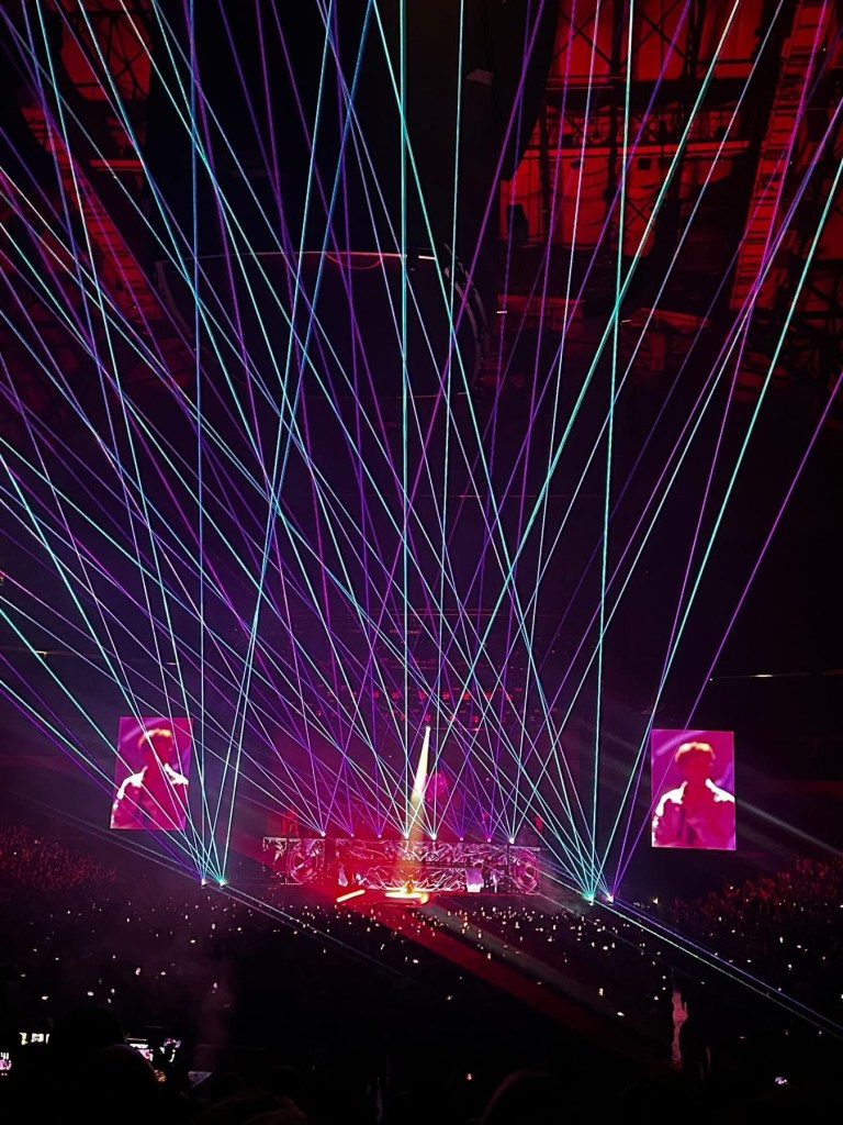 A vibrant concert scene featuring numerous colorful laser lights projecting across the stage, with performers visible on large screens and an audience illuminated by various lights.