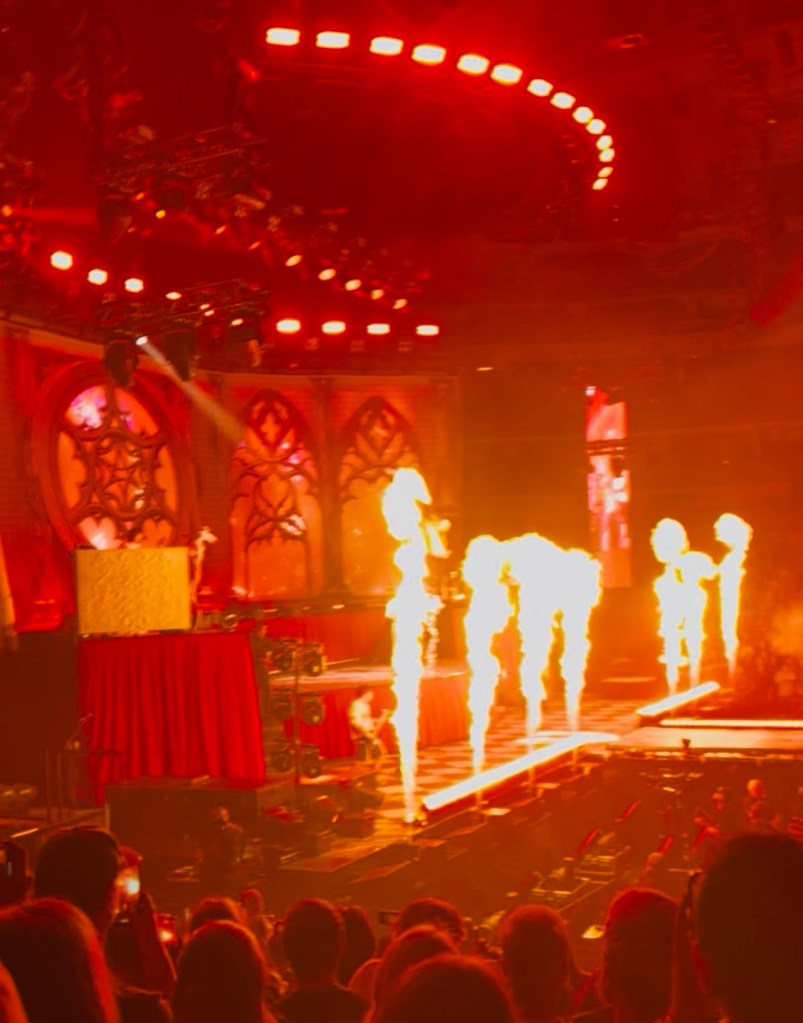 A concert scene featuring a dramatic stage with colorful lighting and flames erupting from the ground, surrounded by an excited audience.