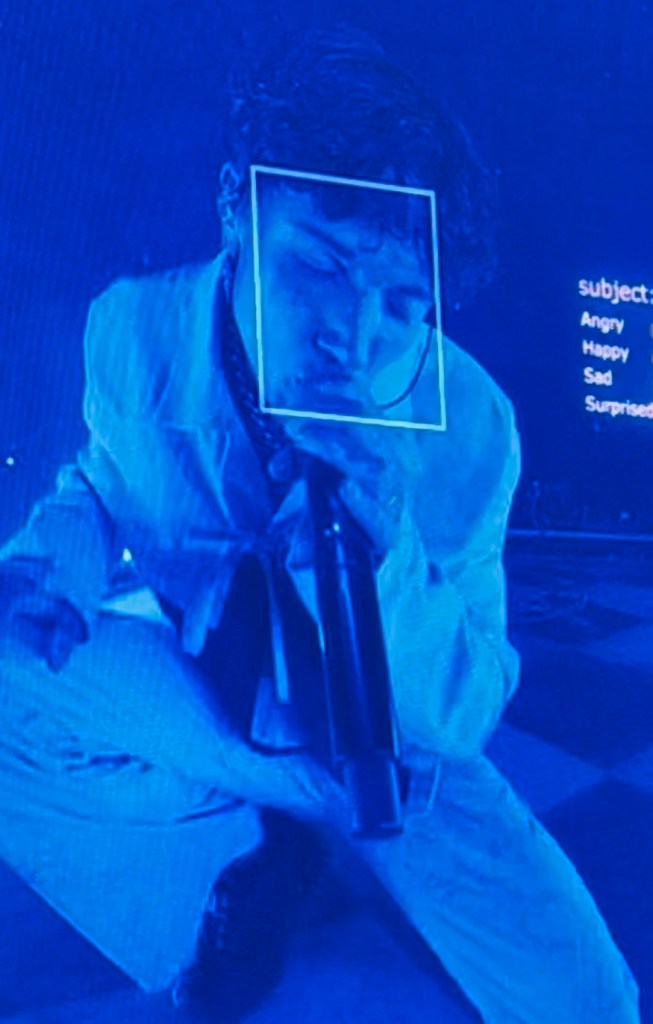 A performer on stage under blue lights, holding a microphone and appearing intense while singing or interacting with the audience.