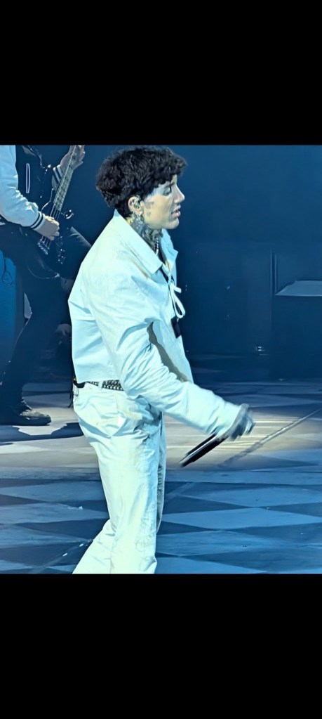 A performer on stage, wearing a white outfit, passionately singing into a microphone during a concert.