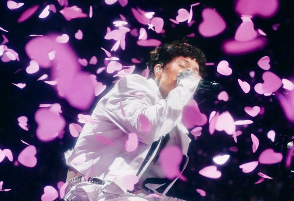 A performer singing on stage surrounded by pink heart-shaped confetti, creating a vibrant and emotional atmosphere.