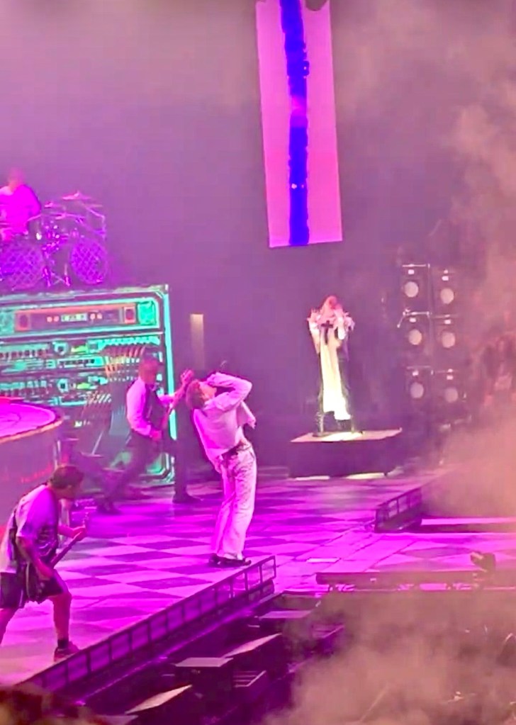 A vibrant concert scene featuring band members performing energetically on stage, surrounded by colorful lights and smoke effects.